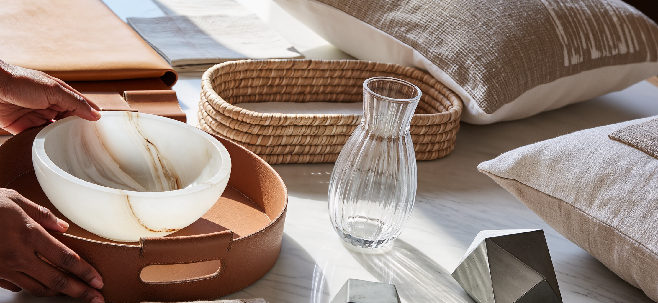 Hands arranging diverse home objects including stone bowl, woven basket, glass pitcher and textiles on marble surface showing editing process